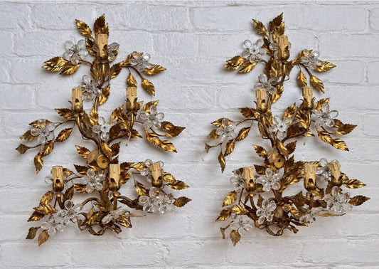 A pair of ornate gold and clear crystal wall sconces with leaf and floral motifs, mounted side-by-side on a white brick wall.